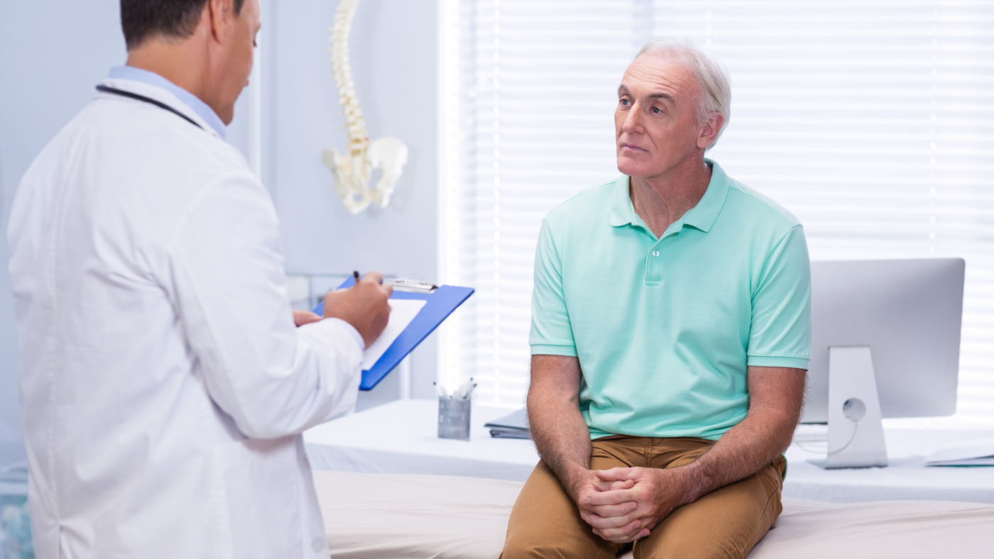 Older man sitting on a medical examination table, attentively listening to a doctor holding a clipboard in a well-lit clinic room.