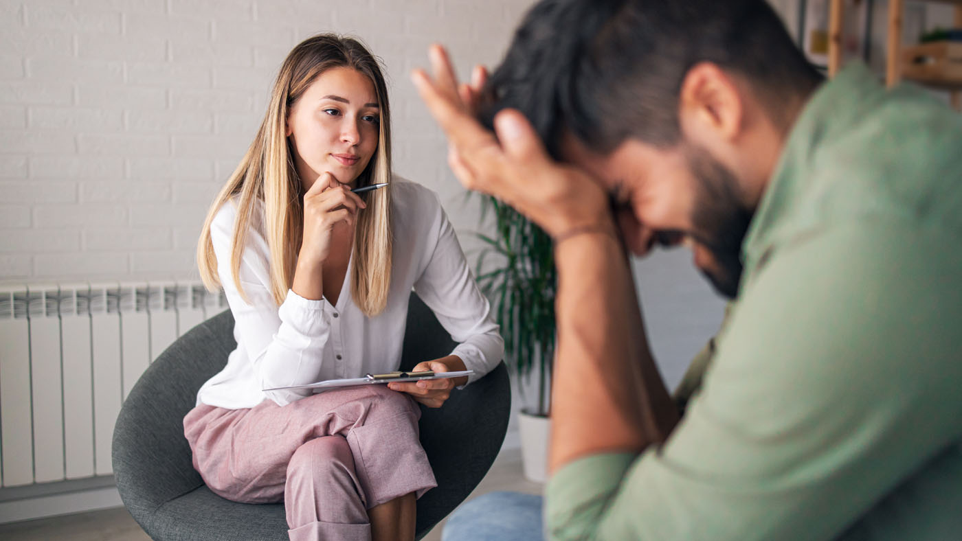 A woman, holding a clipboard, listens attentively to a distressed man with his head in his hands, suggesting a therapy session.