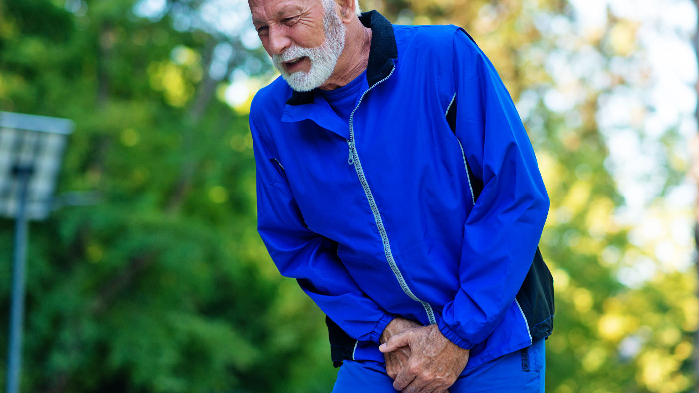 senior man in a blue tracksuit clutching his groin in discomfort, symbolizing urinary incontinence in men.