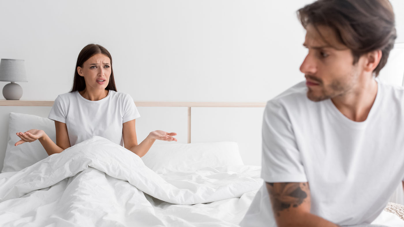 Man sitting on the edge of a bed looking away, while a woman sits in bed looking frustrated, highlighting relationship tension