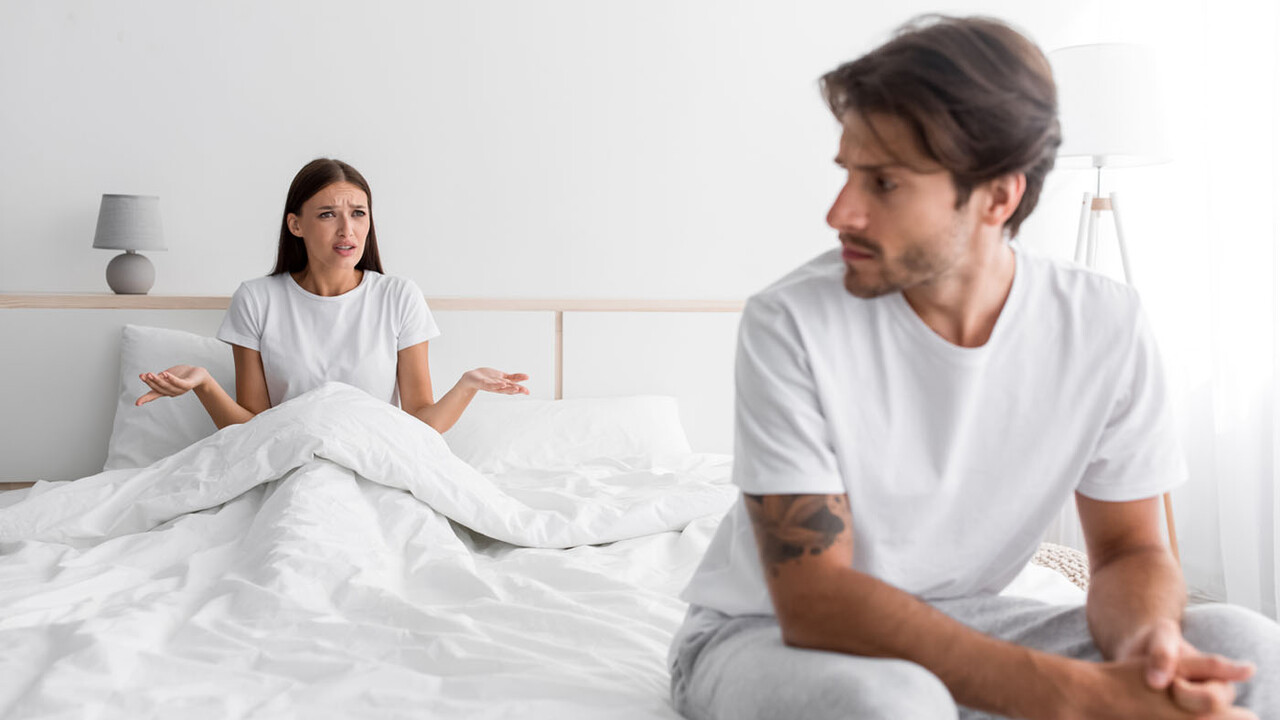 Frustrated couple in bed, woman gesturing while man looks away, illustrating tension related to erectile dysfunction.